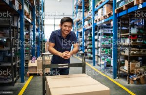 Male worker standing with a trolley with boxes in warehouse. Warehouse worker in uniform with a cart in aisle of storage racks.